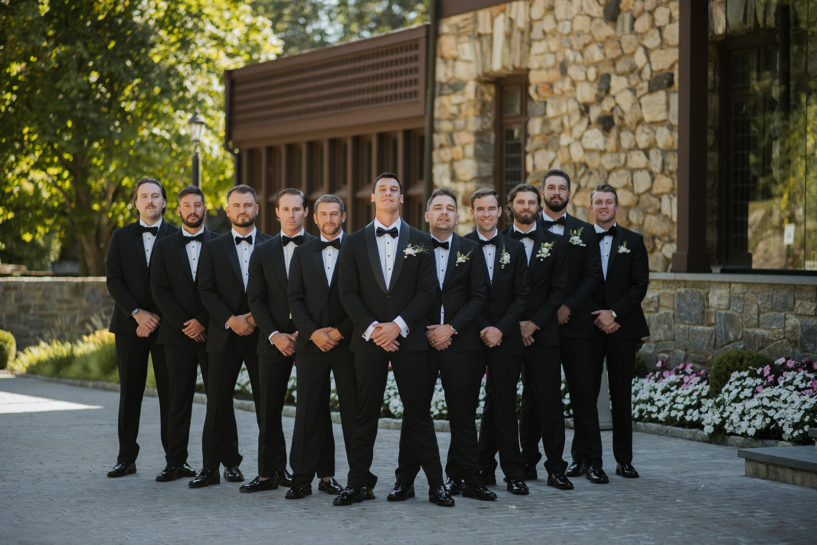 Groom and groomsmen standing in tuxedos, posing proudly outside a historic stone building.